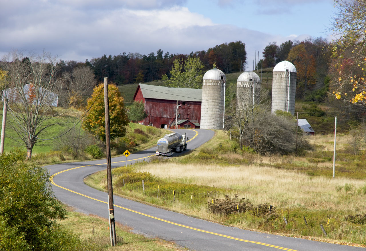 Milk truck driving down two lane highway toward a farm