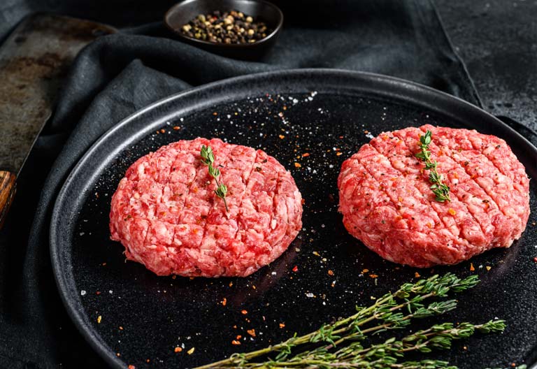 Two raw, seasoned ground beef patties with thyme sprigs on a black plate, with a cleaver and peppercorns in the background.