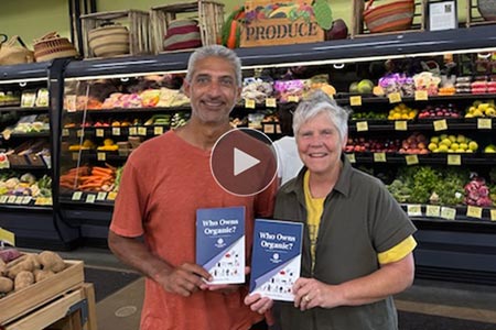 Photo of man and woman in grocery store holding copies of Who Owns Organic? pamphlet