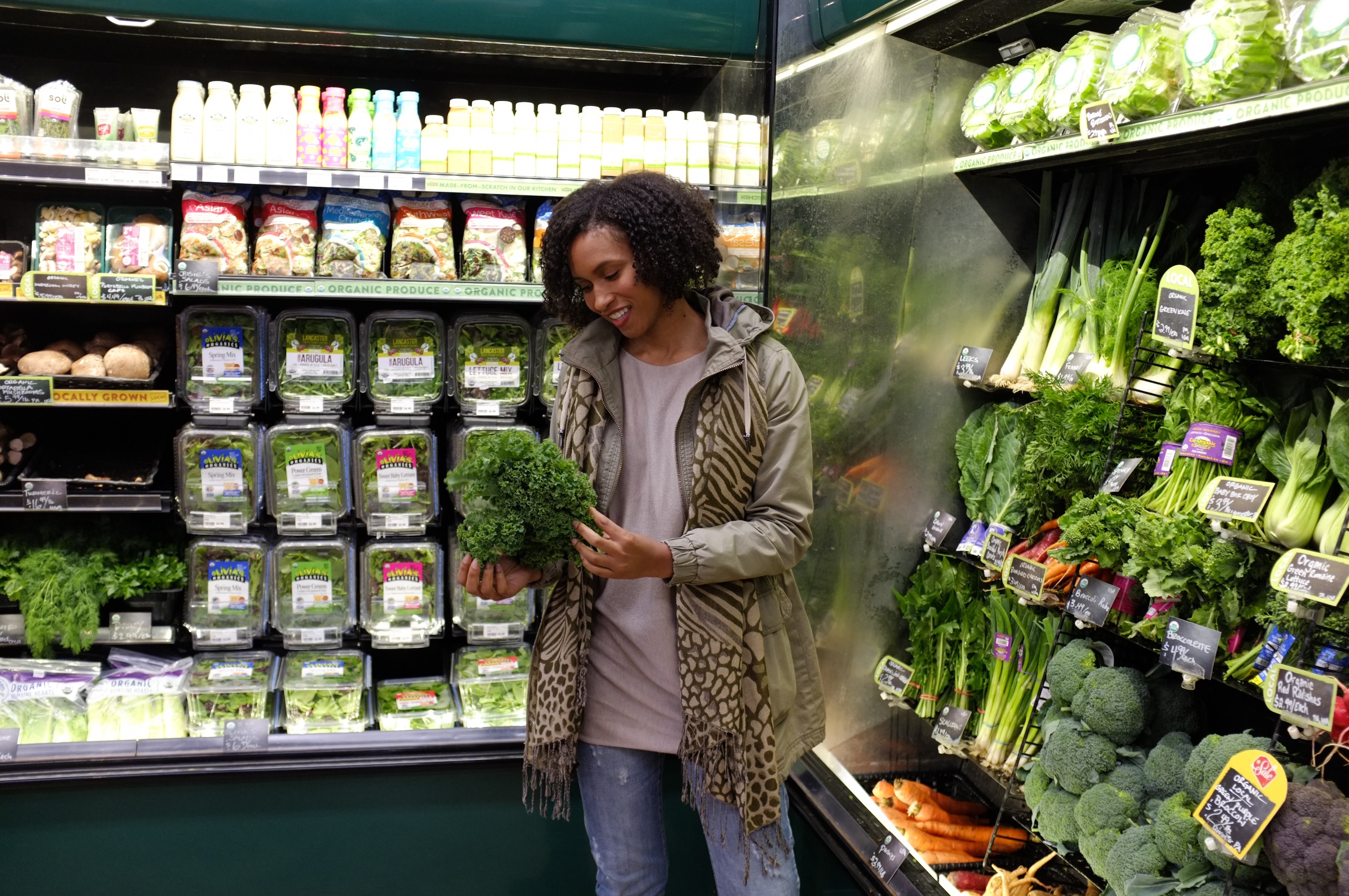 A smiling woman in a grocery store produce aisle holds a bunch of fresh green kale, surrounded by various organic fruits and vegetables.