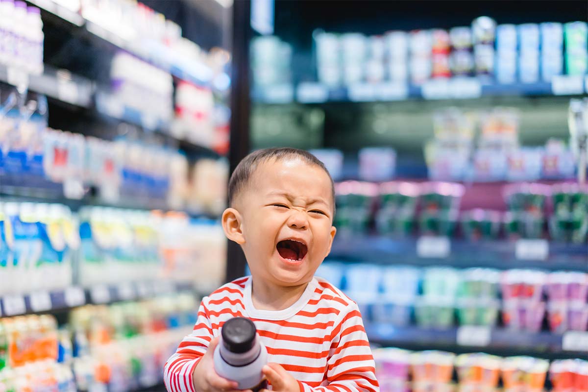 A laughing toddler in a red and white striped shirt holds a bottle in a blurry supermarket aisle.