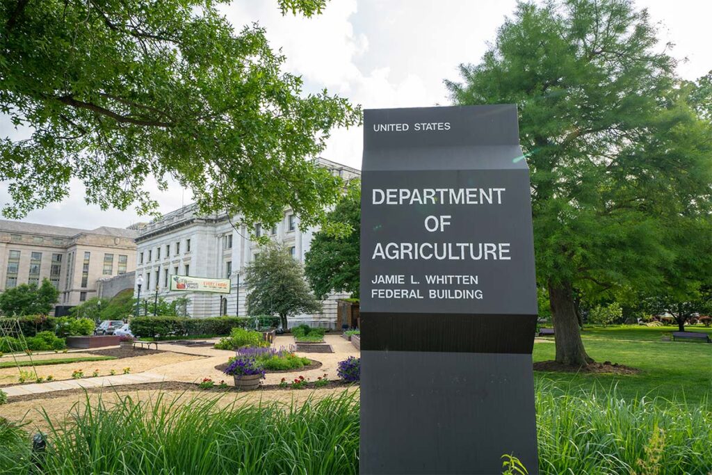 A dark gray sign for the United States Department of Agriculture Jamie L. Whitten Federal Building, with a garden and federal buildings in the background.